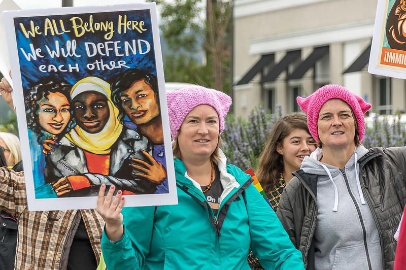A group of women protests. One woman holds a sign with a hand-painted picture of three women holding each other close with the text "We all belong here. We will defend each other."