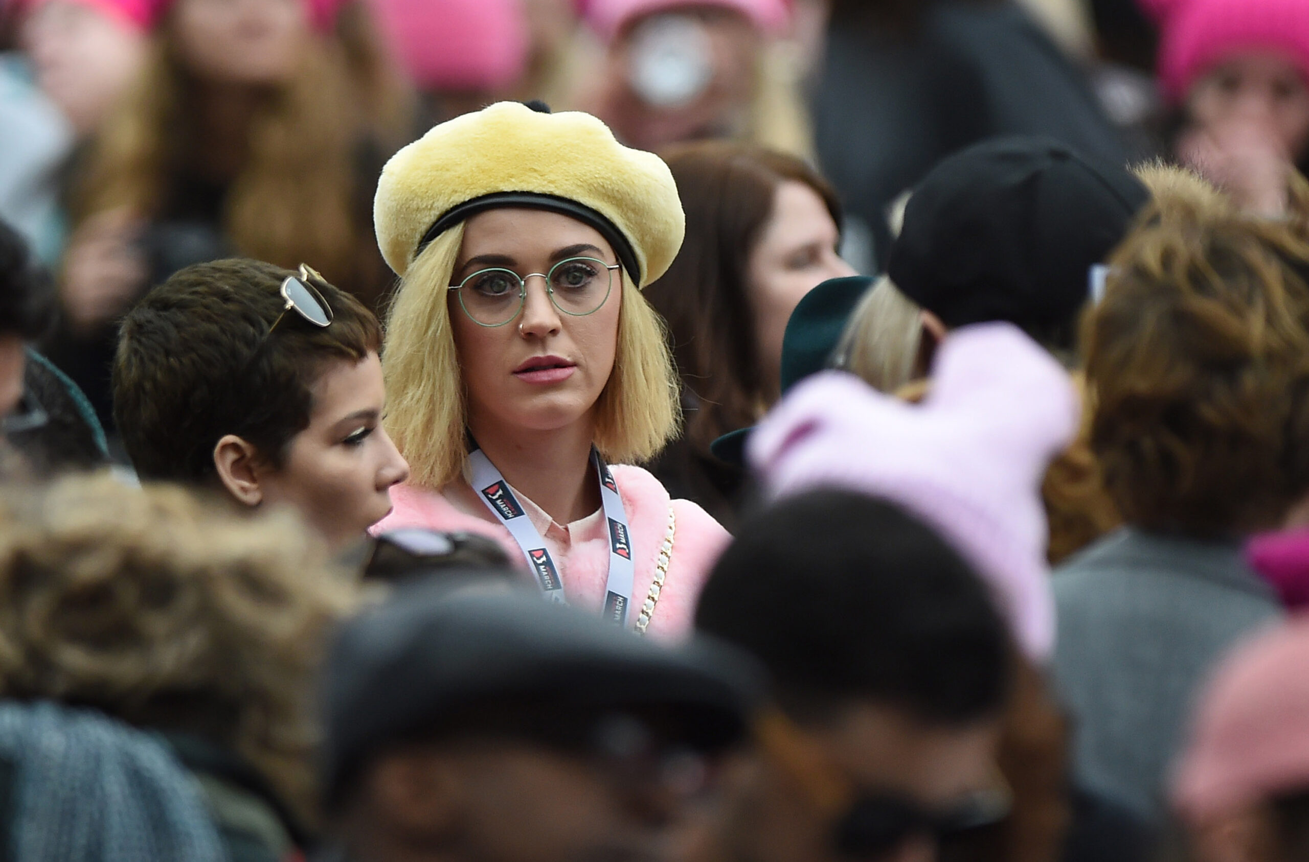 Katy Perry stands amidst a crowd of people, wearing round eyeglasses and a yellow beret.