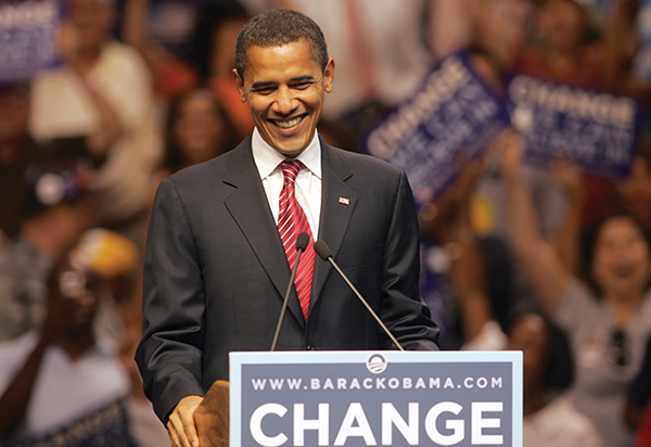 President Barack Obama smiles and stands at a lectern with two microphones at a campaign with supporters (blurred) in the background, holding signs.