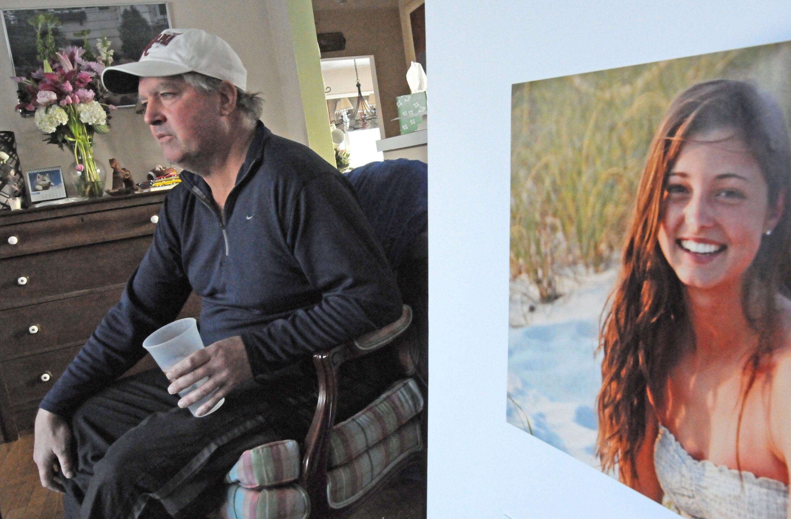 A middle-age man sits with a plastic cup in his left hand and a photo of his deceased daughter on his left.