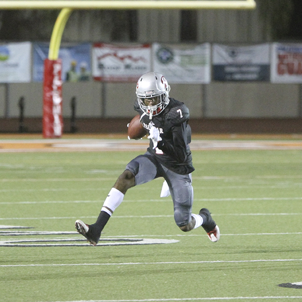 A Palomar football player dashes on a football field, carrying a football in his right arm. He wears a black jersey, gray shorts, and white and gray socks and track shoes.