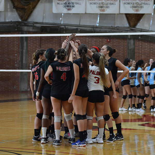 Team players from Palomar's Women's Volleyball gather in a circle and raise their arms up to touch each others' hands. The opposing team in the background gather around on the other side of the net.