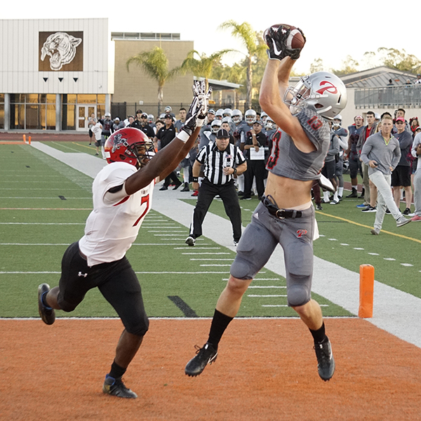 A Palomar football player jumps up and catches a football with both hands as an opposing player tries to intercept with his both hands. A referee and a crowd of Palomar players and coaches stand in the background and watch them.