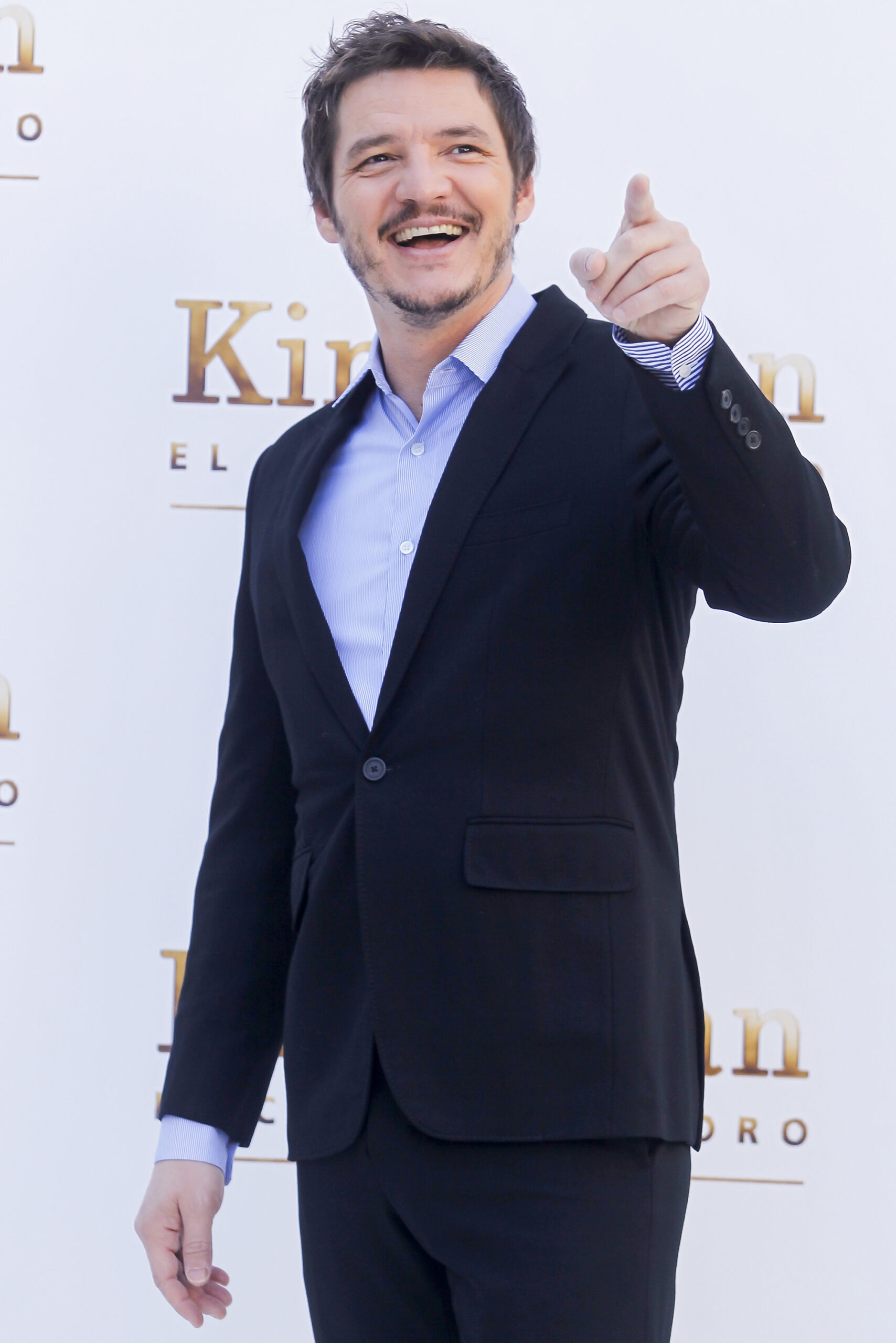 Actor Pedro Pascal smiles and points in front of him, wearing a dark blue suit and a light blue shirt.