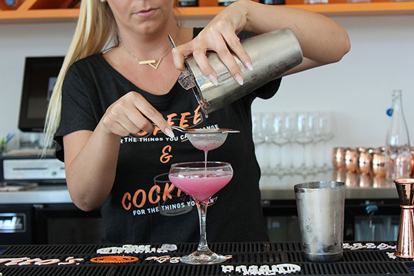 A female barista pours a pink liquid through a strainer held in her left hand. The liquid fills a large, martini-like glass.