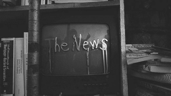 Black and white photo of a cathode TV in a bookcase with the words "The News" painted on the screen with a paint-like substance. A reflection of a person giving someone a middle finger is in the middle of the screen.