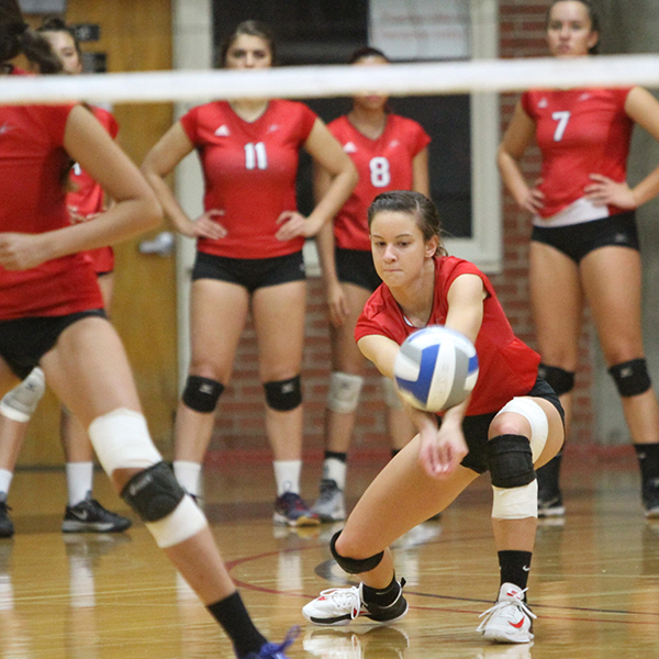 A female volleyball player bumps the ball with her forearms. as her teammates watches her in the background.