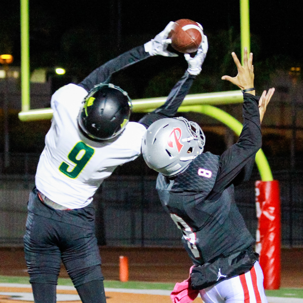 A Palomar football player (right) reaches his hands up to catch the football, but an opposing player had jumped up and caught it with both hands. A yellow field goal is in the background.