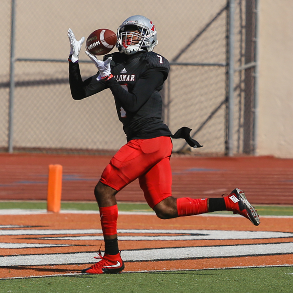 A Palomar football player catches a football with both hands in front of his face as he runs. He wears a black jersey, red game shorts, red and black socks, and red shoes.