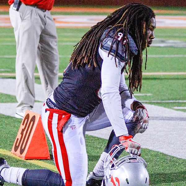 A Palomar football player kneels on the ground on his right knee, holding his helmet at the chinguard with his right hand with the helmet on the ground. He sports long, black dreads.
