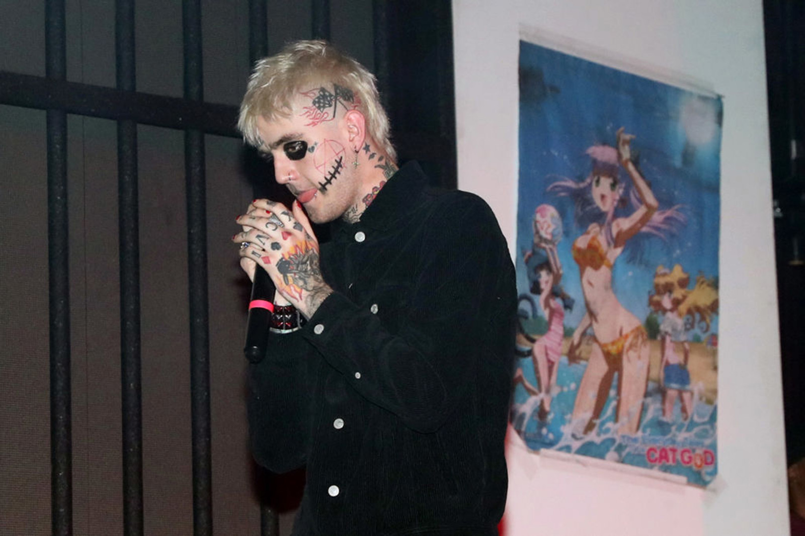 Lil Peep performs with both hands on a microphone with an anime poster behind him. He has tattoos on his hands and face, sporting a black buttoned-up coat.