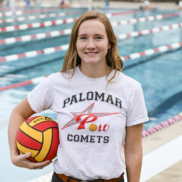 Palomar water polo player Sydney Thomas stands by a swimming pooling, holding a water polo ball in her right arm against her hip.