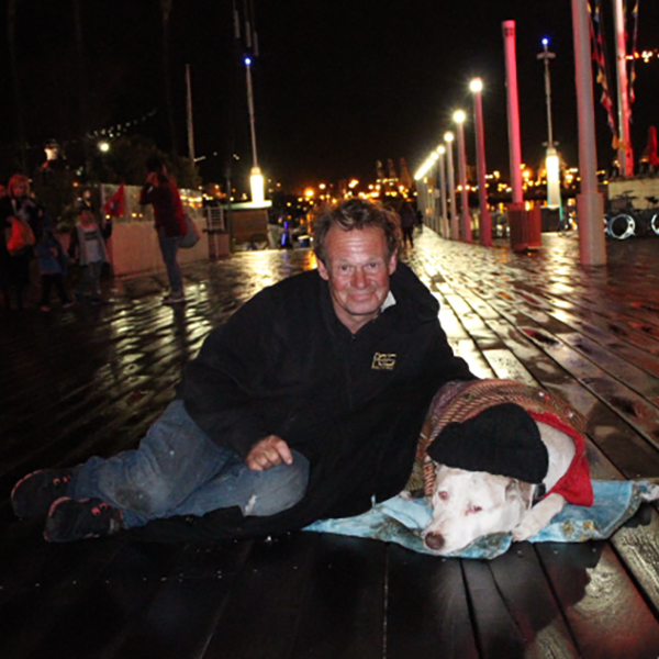 An older homeless man lies on a boardwalk with his dog, looking at the viewer. The dog wears a black beanie and is covered with a brown blanket. The man wears a black hoodie and jeans. Several people stand or walk in the background.