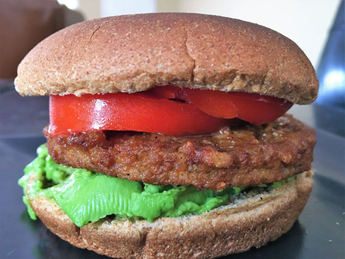 A vegan burger with avocado and tomato slices, and a vegan patty between two wheat burger buns.