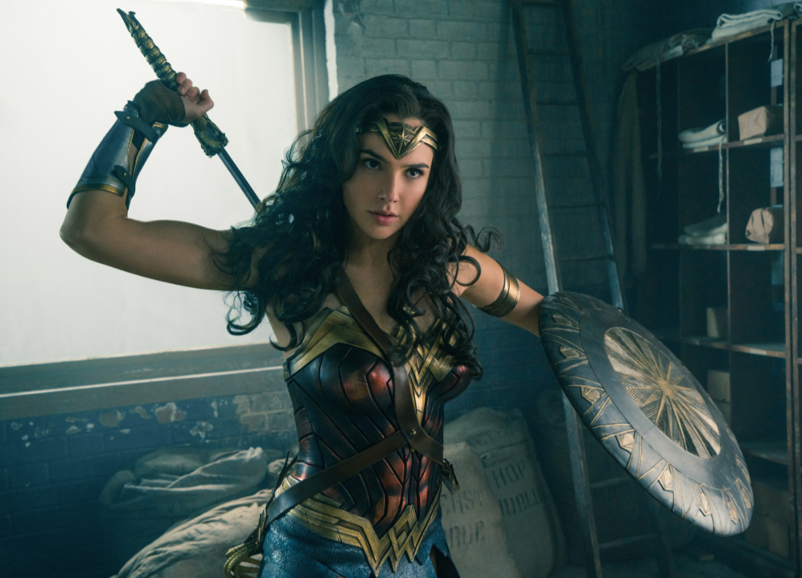 Wonder Woman, played by Gal Gadot, unsheathes her sword behind her back while carrying a shield in her left arm.