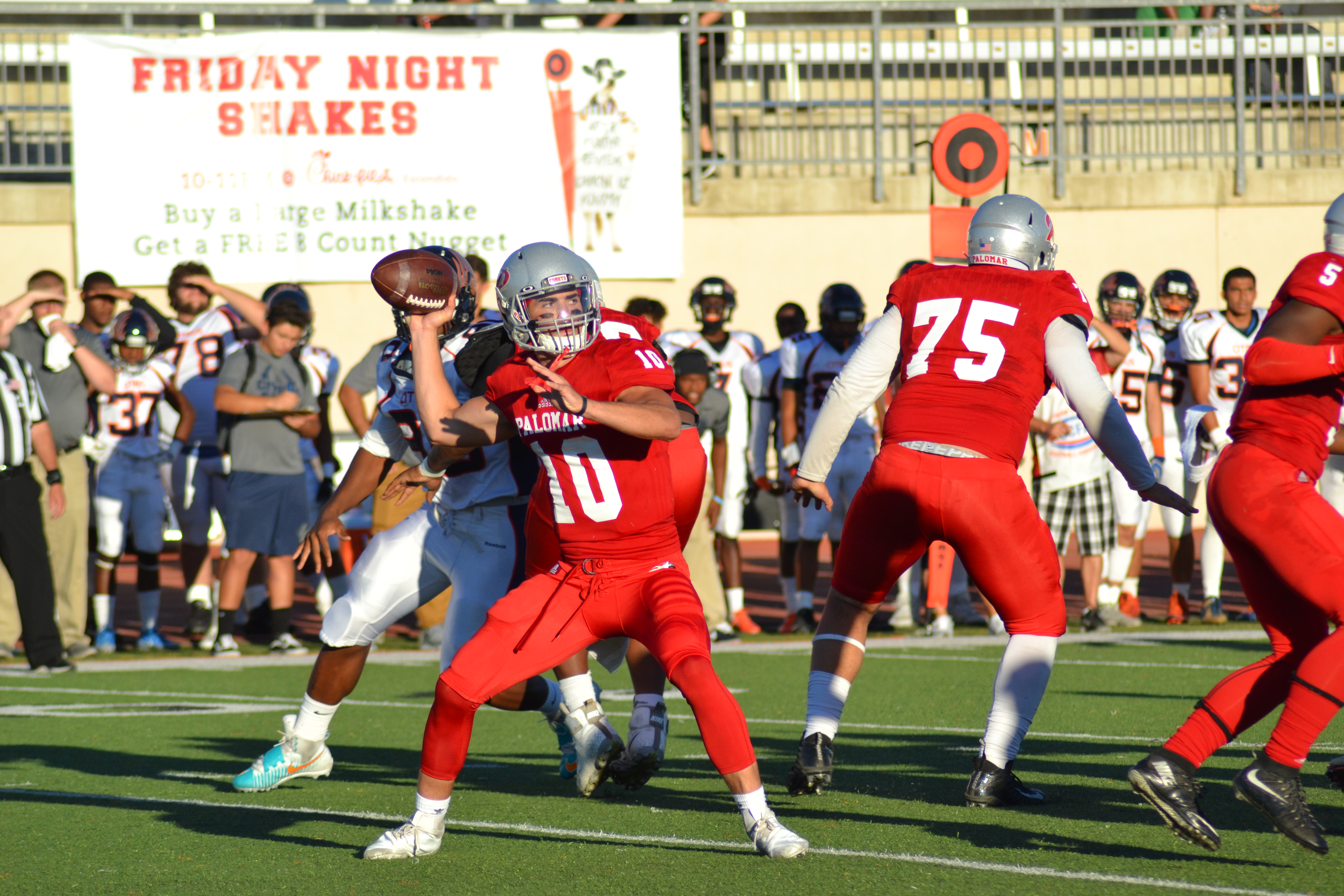 A Palomar football player in a red uniform and gray helmet brings his right hand behind his head to throw a football as other players tacklet in the background. A group of players, coaches, and a referee stand in the background.