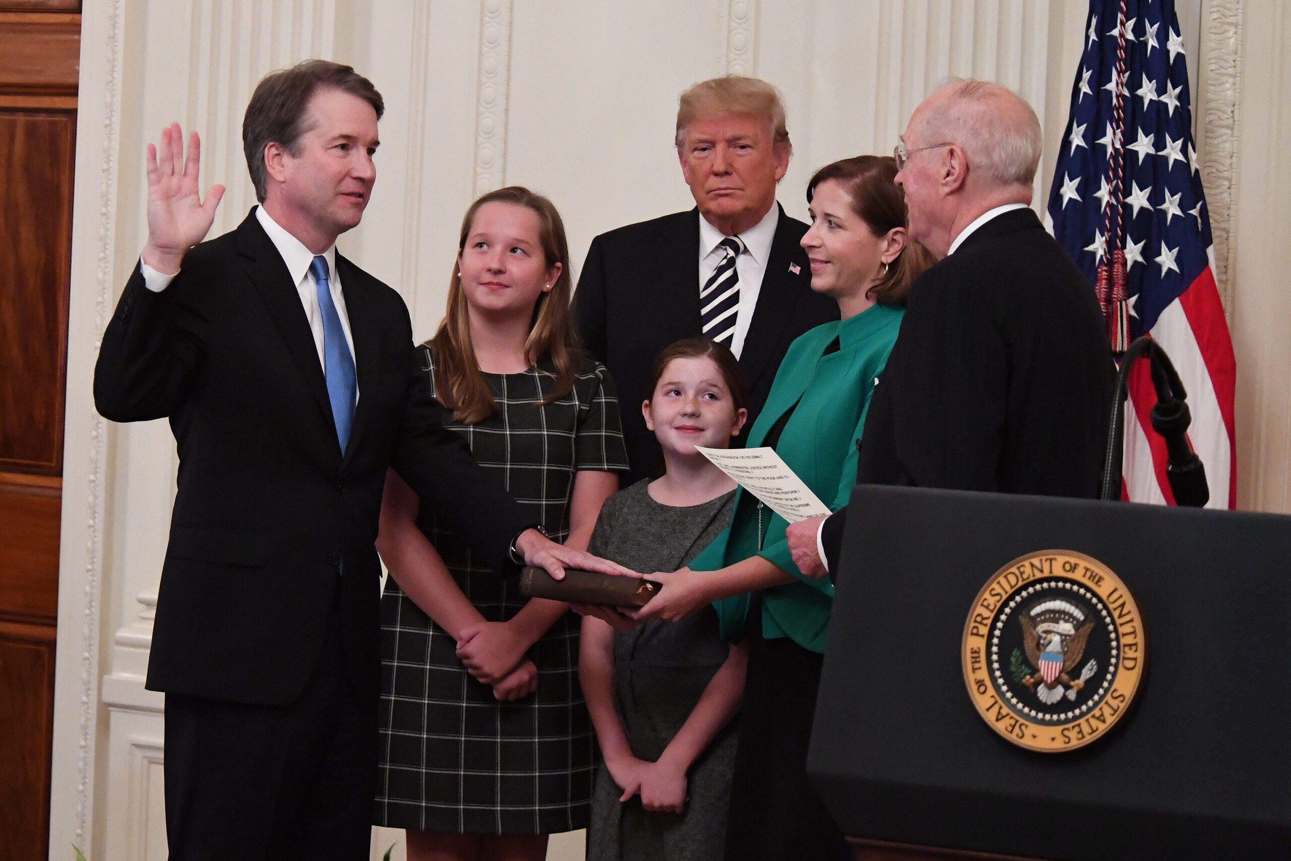 Brett Kavanaugh raise his right hand and put his left hand on a book as he swears an oath. His two daughters and wife, President Donald Trump and Justice A. Kennedy looks on.