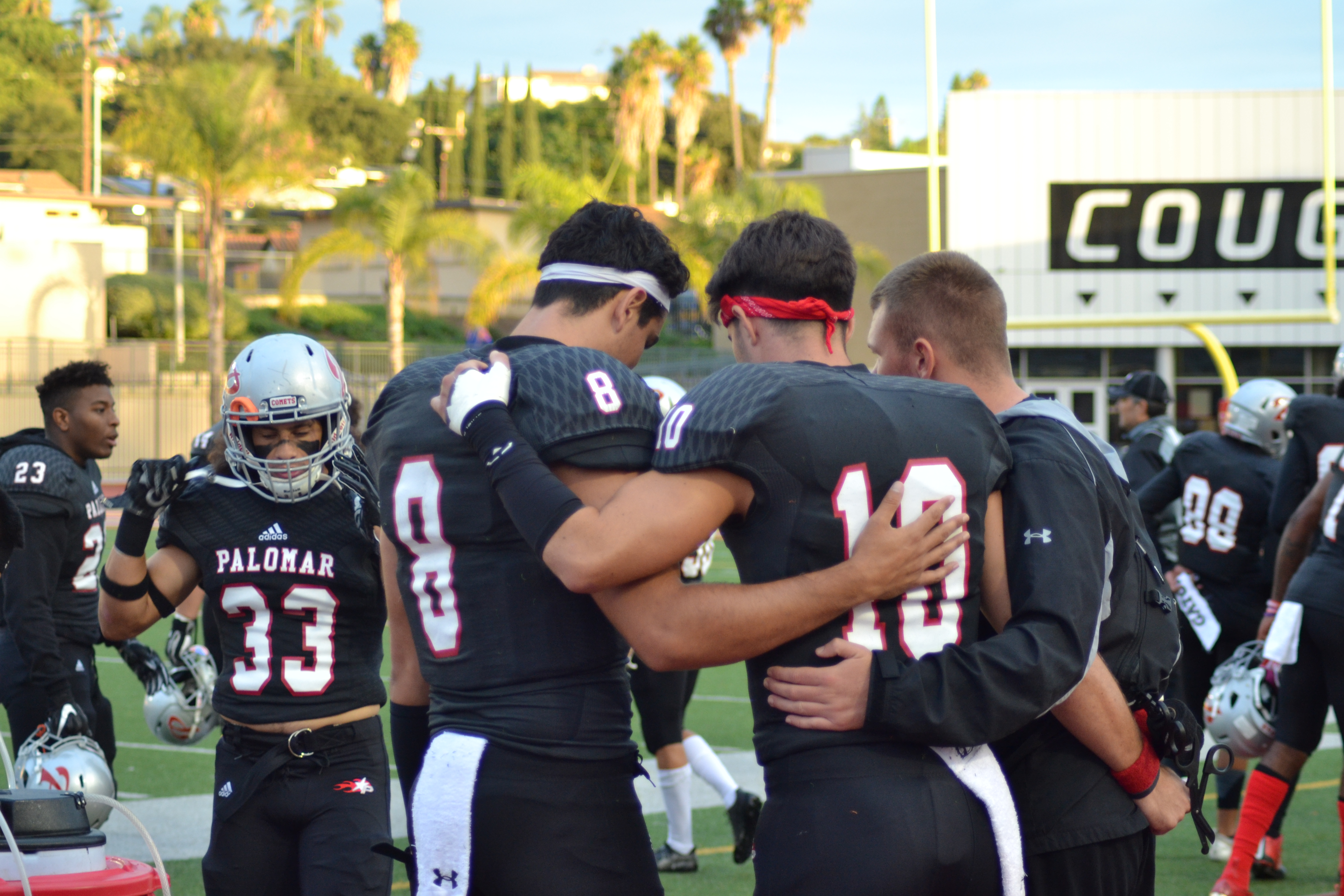 Three Palomar football players huddle with their arms around each others' back and waist. Several players mill around them on the field.