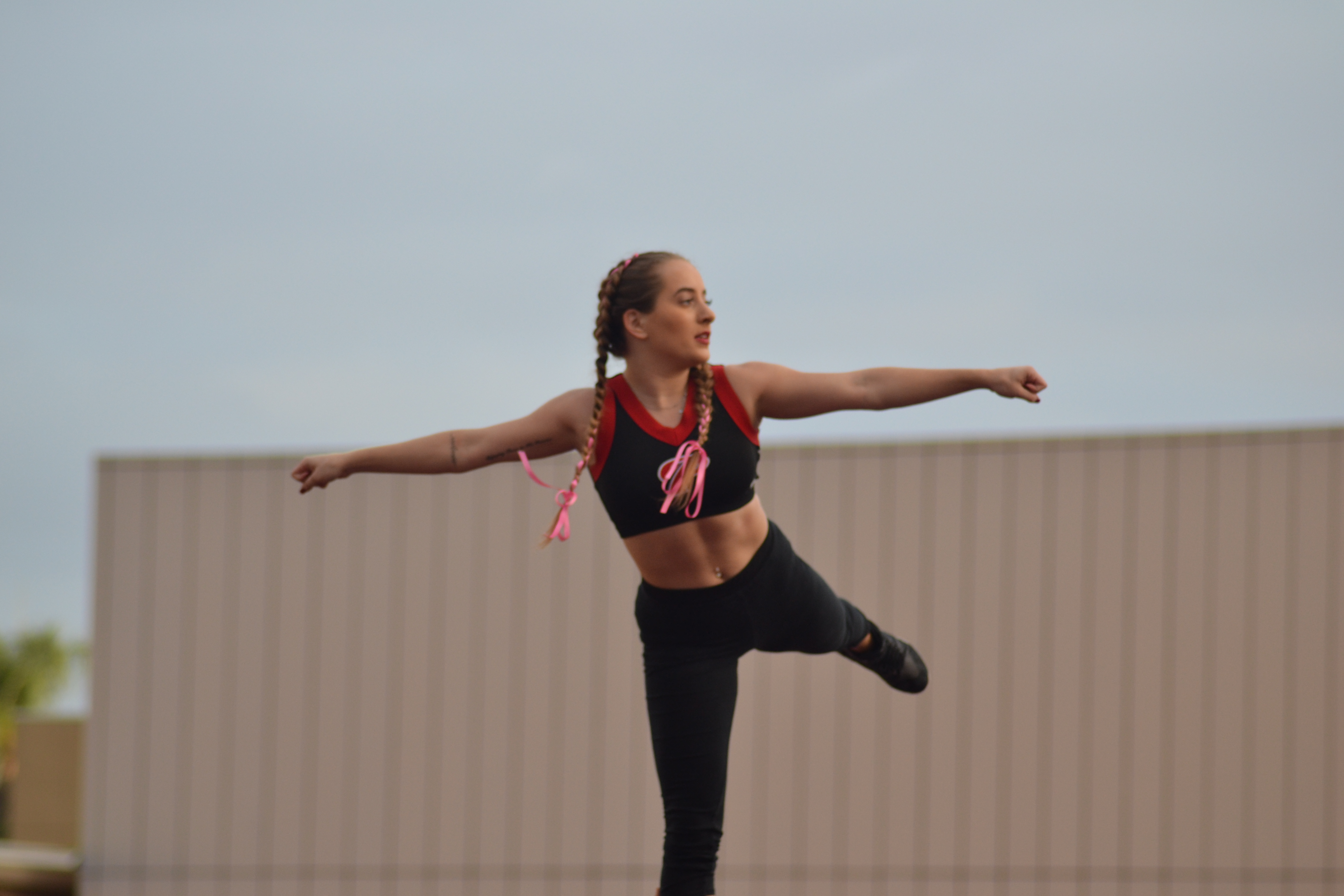 A female cheerleader in a black top and black pants and shoes stand on her right leg, stretches both arms to her sides in a fist, an extends her left leg behind her. She looks to the right.