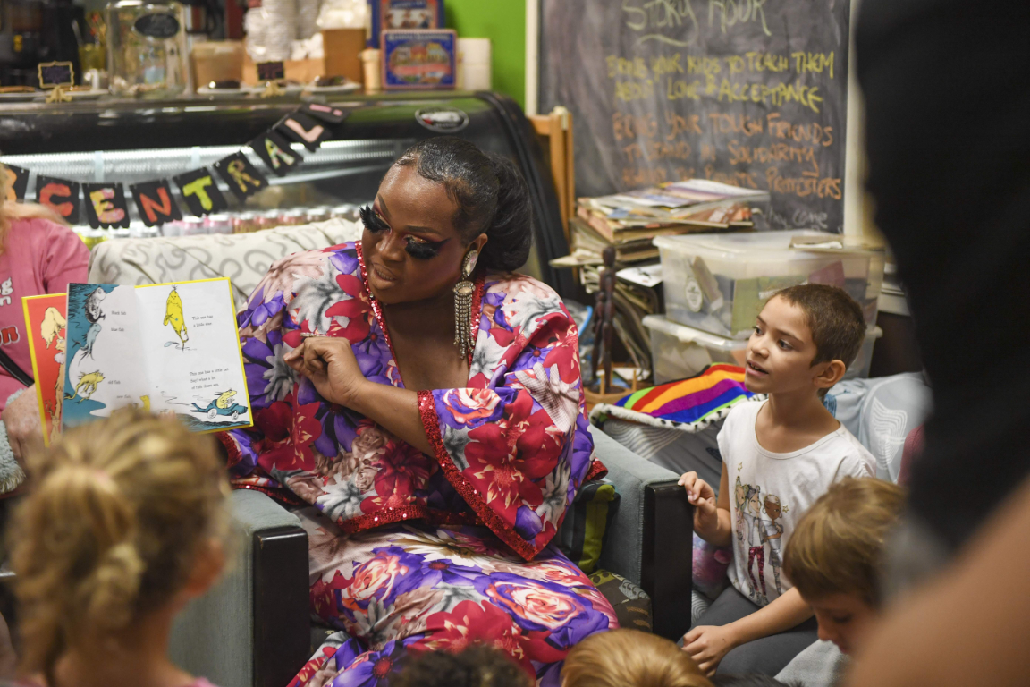 A drag queen holds a Dr. Seuss book with several children gather around her in a library.