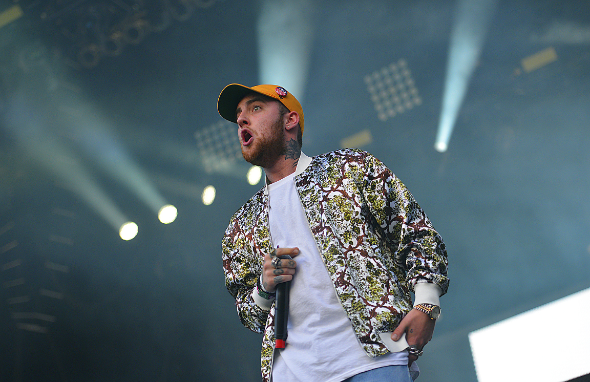 Mac Miller performs on a stage, holding a microphone in his right hand and sporting an orange cap and a patterned jacket.