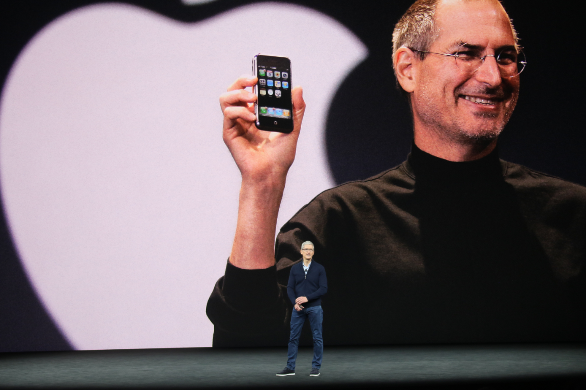 Apple CEO Tim Cook stands on a stage with a huge image of Steve Jobs holding a smartphone in the background.