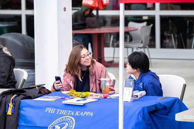 Two young women talk to each other a table with a blue covering that says "PHI THETA KAPPA."