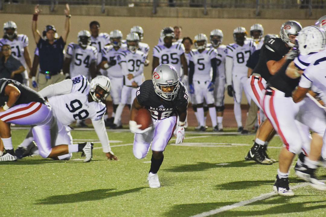 A Palomar football player runs and ducks behind a group of other players who are tackling each other. He holds a football in his right hand. A group of players and a referee stands in the background.