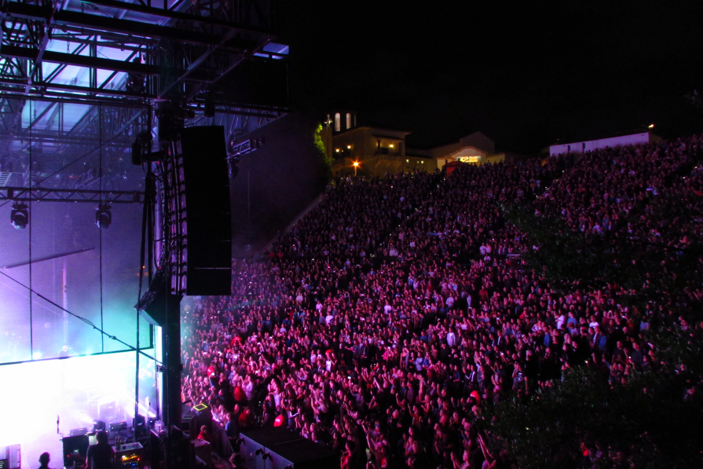 Thousands of people gather at an amphitheater to watch a rock band playing at night.