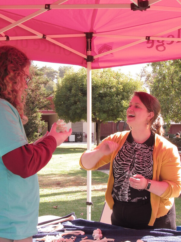 A female professior wearing an orange sweater and black shirt with a white outline of a skeleton talks to a man with long read hair who is holding a bone mold beneath a red tent.