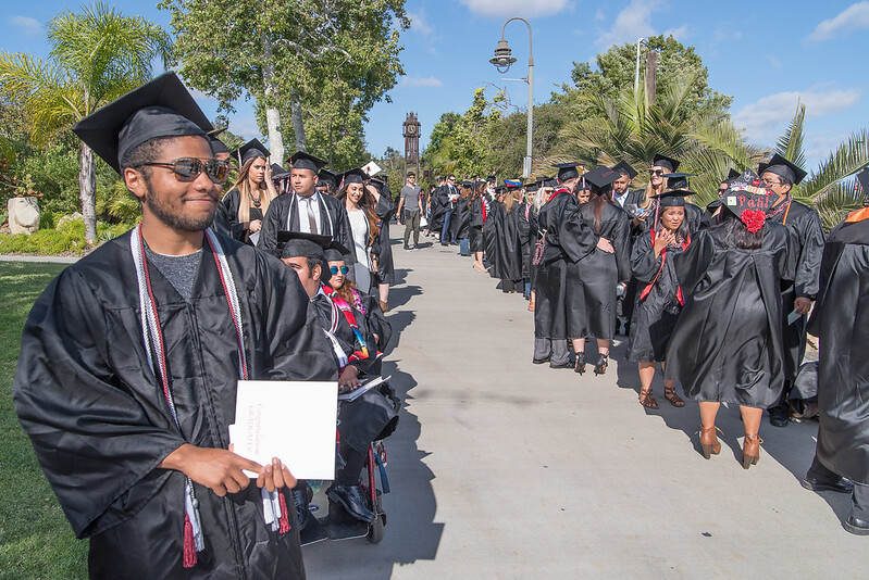 Many students stand or sit at a Palomar College graduation ceremony along a walkway that leads to the clock tower.