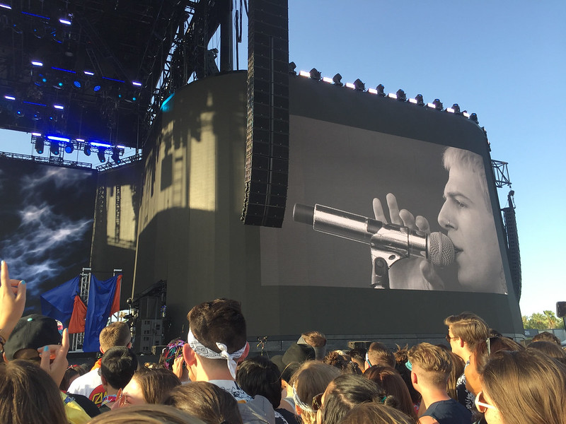 A crowd gathers by a stage at Coachella with a big screen showing a man singing at a microphone.