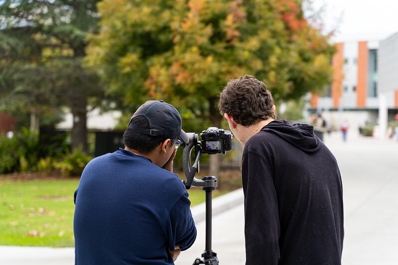 Two male students look through a digial camera lens on a tripod.
