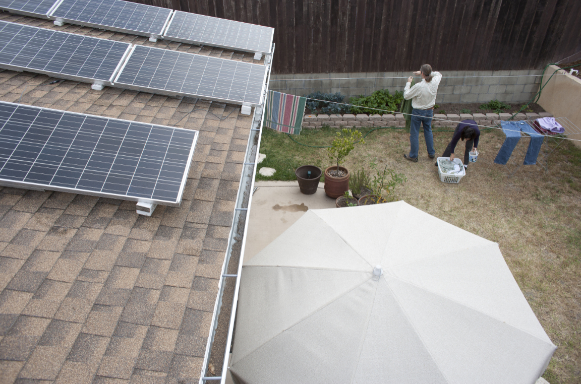 A man and a woman hang laundry in a backyard. Several solar panels lie on a roof to the left, and a white unbrella in at the bottom.