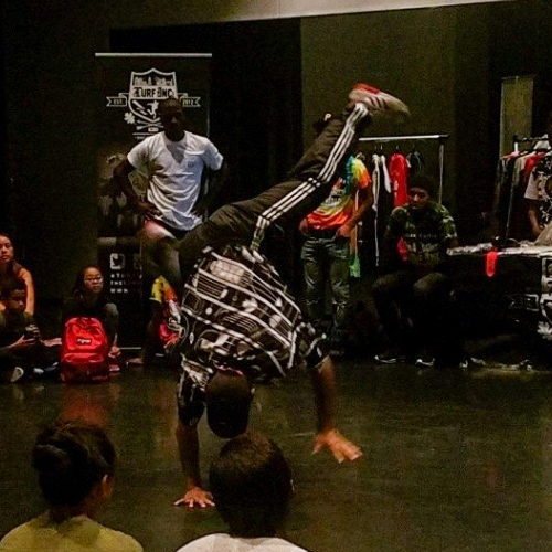 A male breakdancer spins on the ground on his hands with his legs flying in the air like a windmill. Several students sit or stand in the background and foreground watching him.