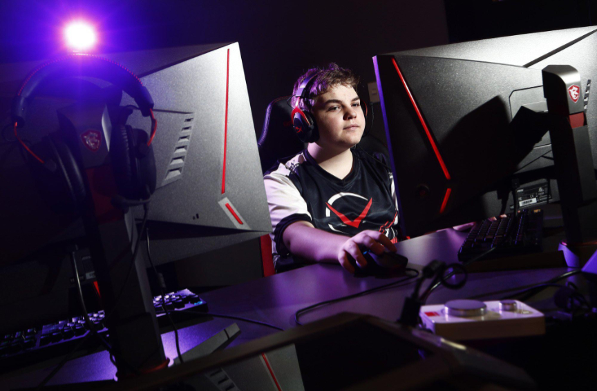 A male teenager plays at a desktop computer wearing black and red headphones. A purple spotlight shines from the upper left.