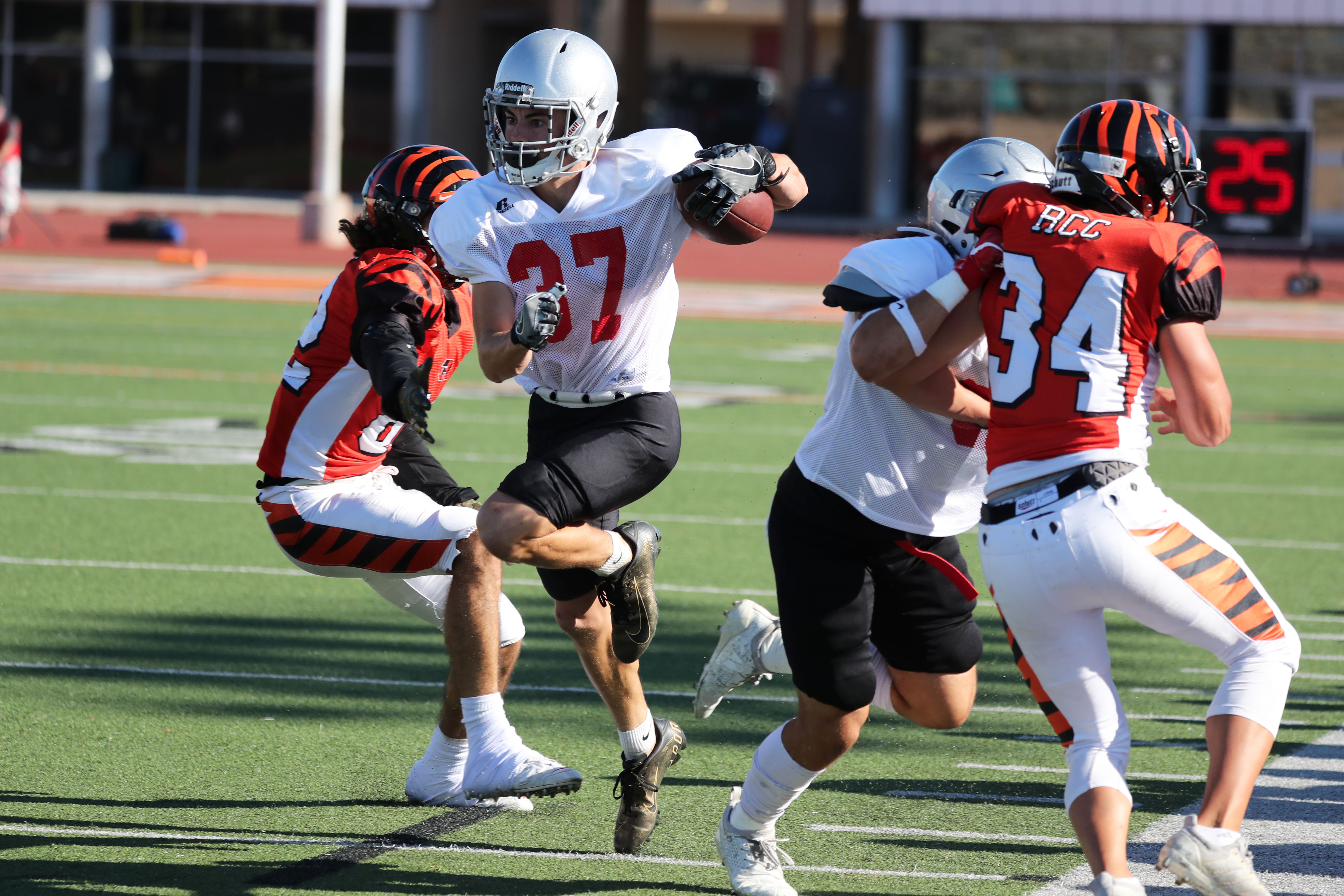 A Palomar football player (37) runs pass an opposing player, carrying a football in his left arm. A team player and an opposing player tackle each other on the right.