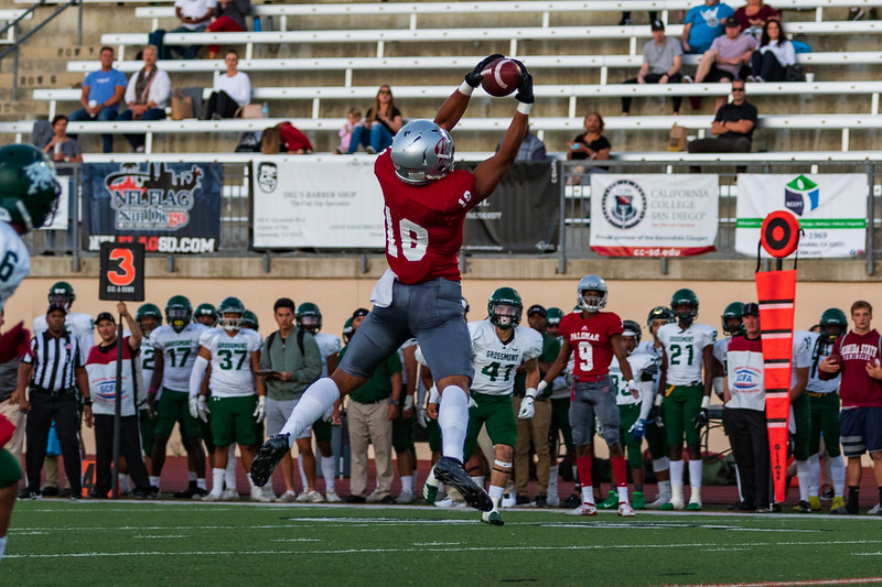 A Palomar football player jumps up and catches a football with both hands. A crowd of other players and coaches stand in the background. A small crowd of spectators sit on the bleachers in the background above them.