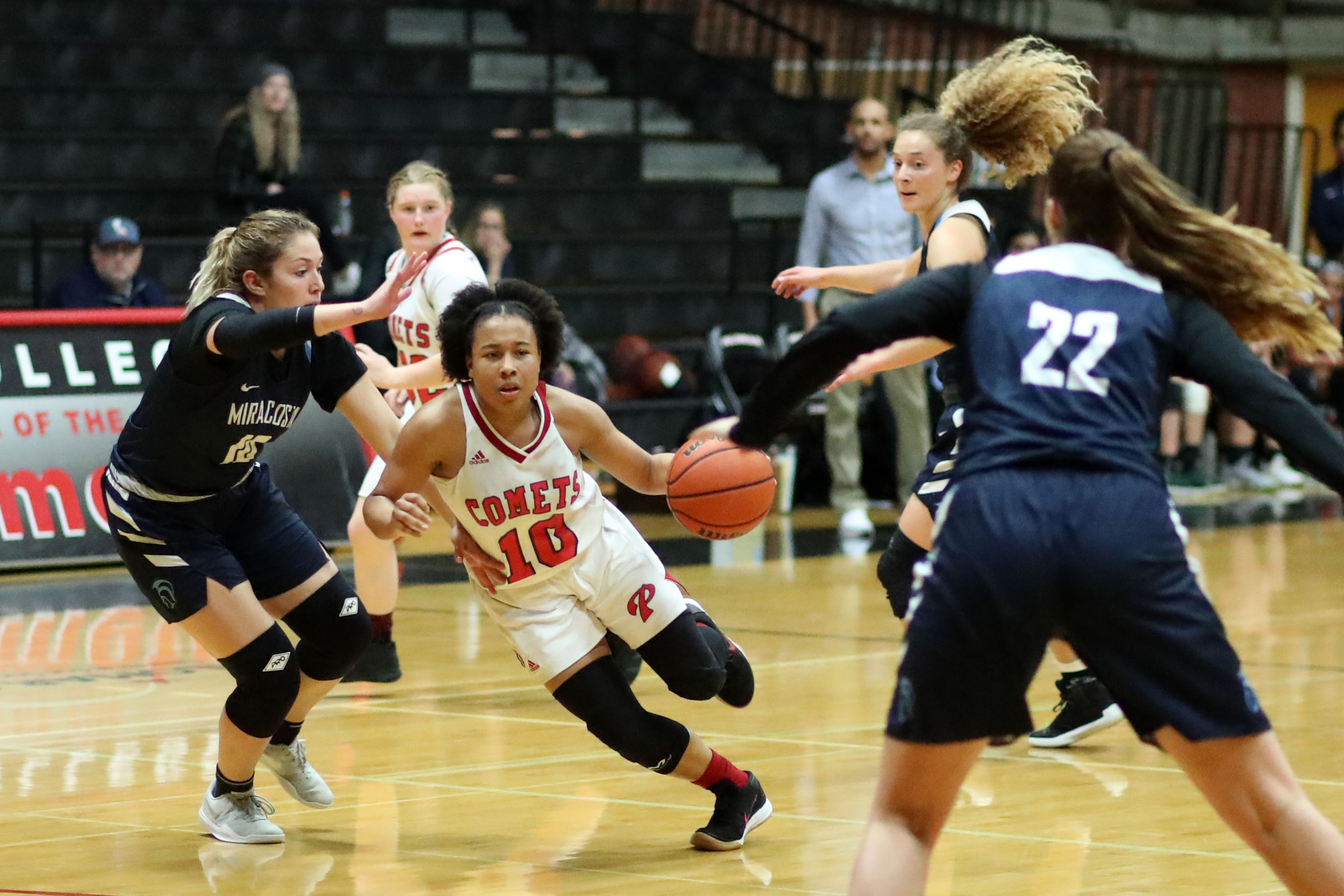 A female Palomar basketball player runs between three opposing players while dribbling a basketball in her left hand.