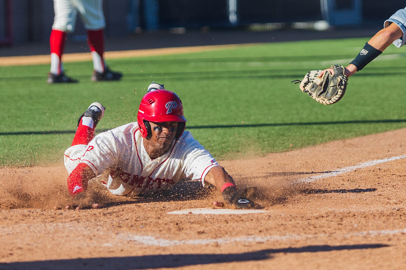 A male Palomar baseball player slides toward a base on his stomach. A arm with a mitt extends toward him on the upper right.