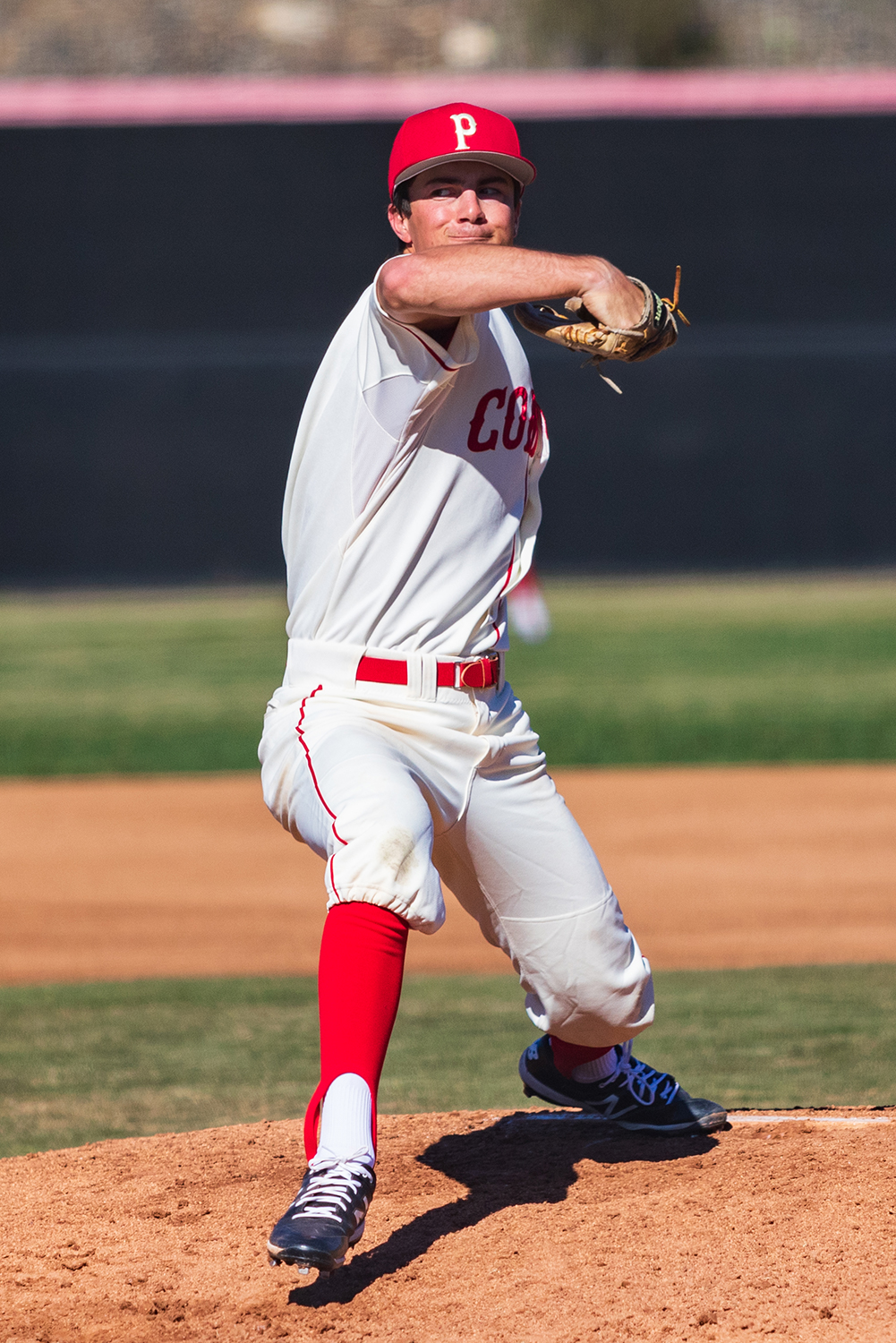 A male Palomar baseball pitcher prepares to throw a baseball with his left hand, which is hidden behind his body due to a direct front view of the pitcher.