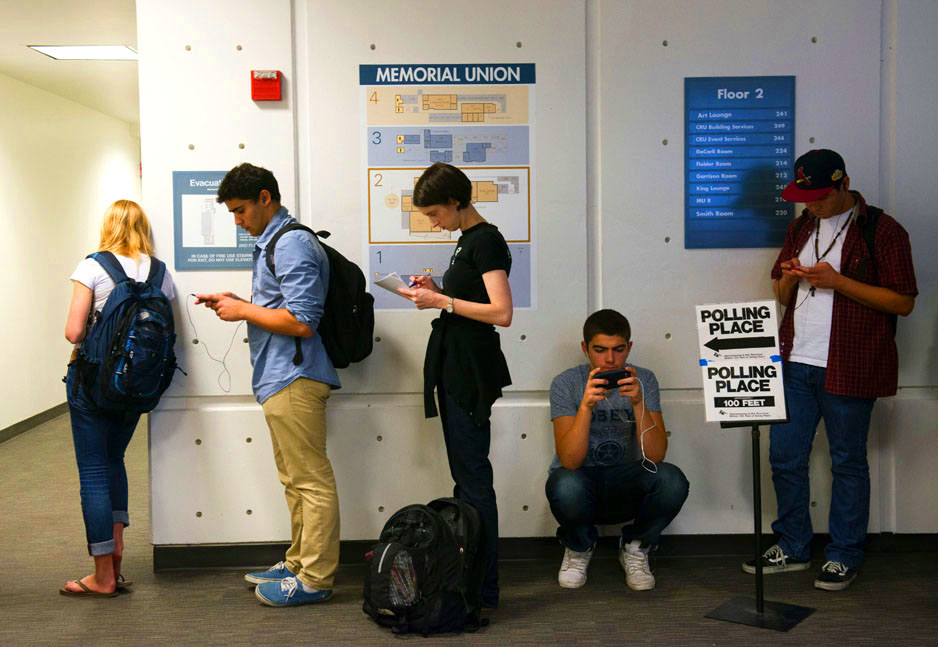 Five students stand in line waiting to vote. One of them is squatting and all but one are looking at their smartphone.