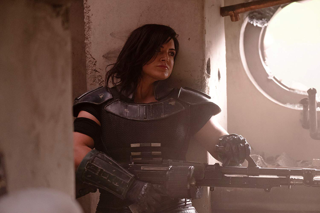 Actress Gina Carno as Cara Dune in the first season of "The Mandalorian." She wears black body armor and wields a large rail gun as she peers out of a round window.