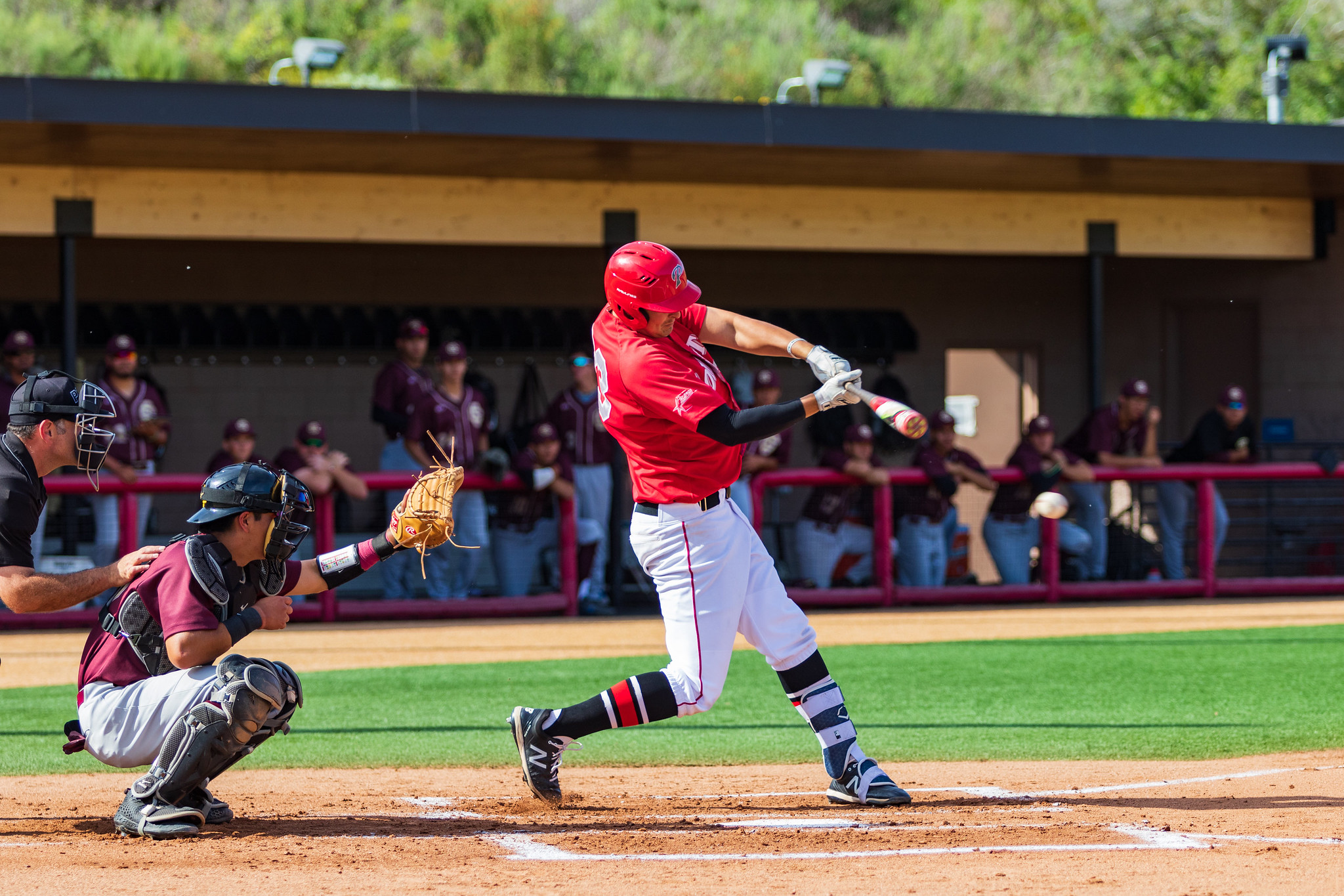 A male Palomar baseball player wearing a red jersey and white pants swings a baseball bat. A catcher squats behind him with his mitted left hand raised, while an umpire watches from behind. More than a dozen opposing team players watch from the dugout (blurry).