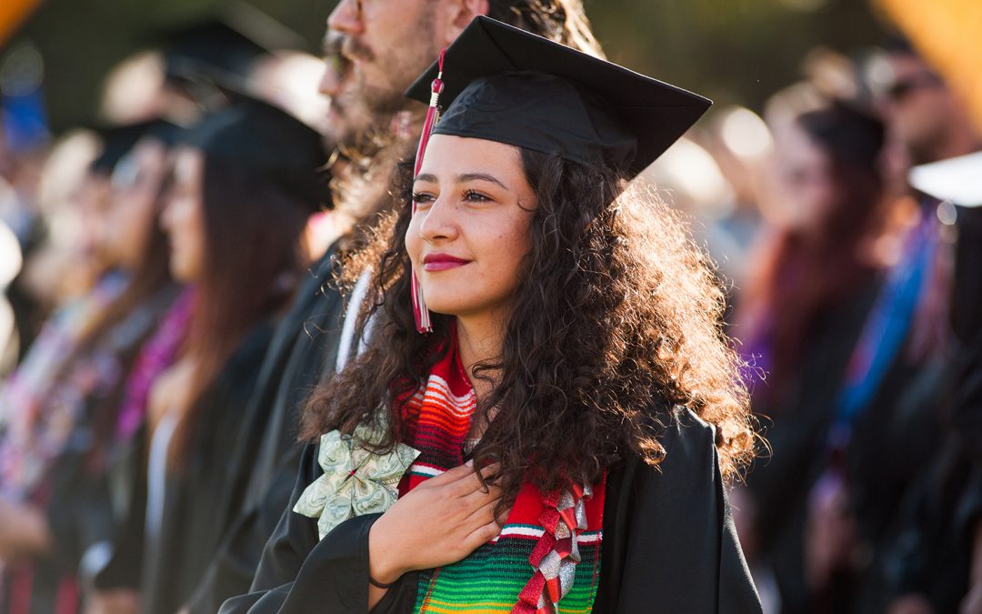 A young Hispanic woman wears a black cap and gown for Palomar College's graduation ceremony. She wear a large red, green, and white scarf that drapes around her neck, resembling a Mexican flag pattern. She has her right hand on her chest.