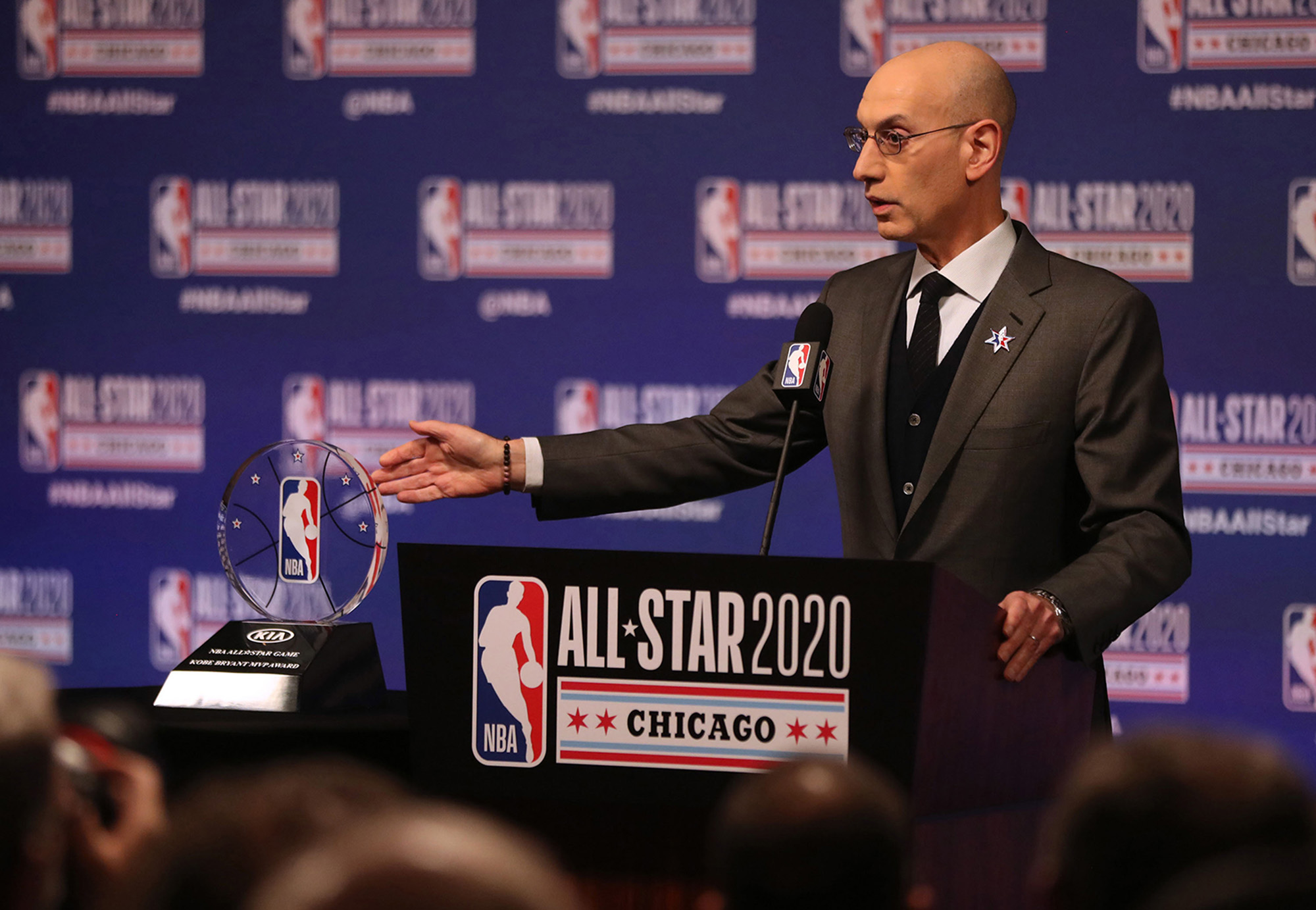 NBA Commissioner Adam Silver talks at a lectern with a crowd in front of him. He gestures with his right hand toward an NBA trophy. The lection has an NBA logo to the left and the text "ALL STAR 2020 Chicago" next to it.