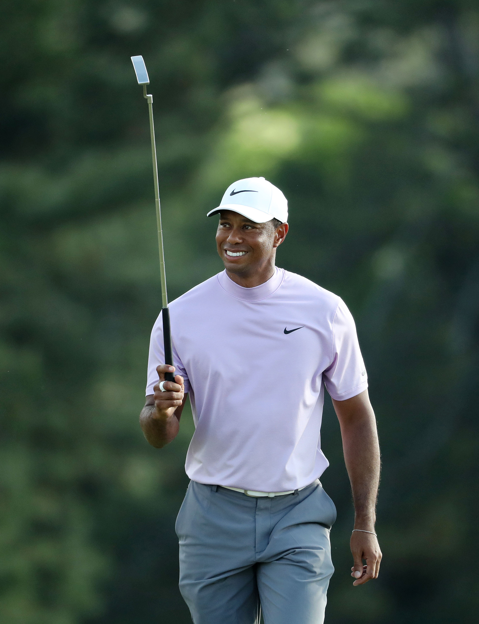 Tiger Woods walks and holds a golf club vertically up with his right hand. He wears a white cap with a Nike logo, a light pink shirt with a Nike logo, and light pastel blue-green pants.