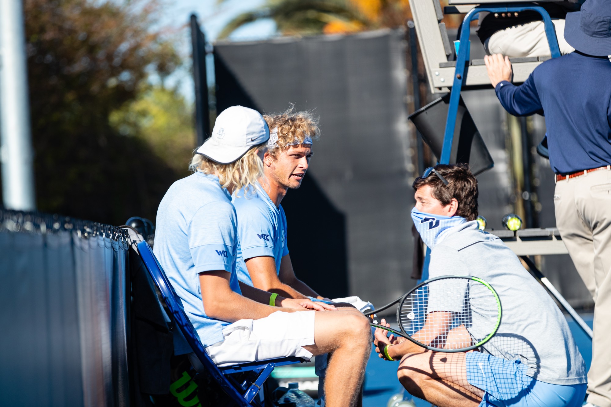 Coach Ryan Keckley speaking to two men's tennis players during a home match against Cal Poly San Luis Obispo.