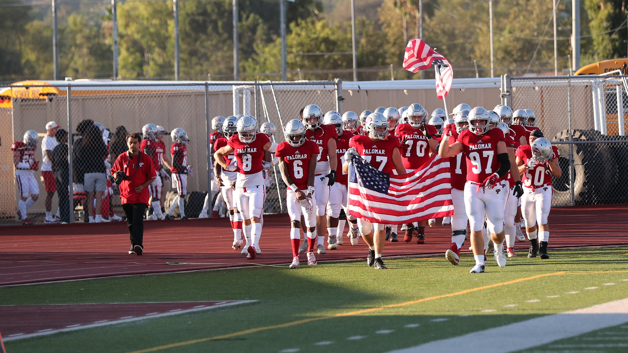 A group of Palomar football players run on a field. Two of the players in the front carries an American flag.