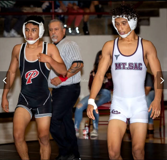 Two wrestlers—one from Palomar College (l) and one from Mt. Sac—stand near each other before a match. A referee walks behind them.